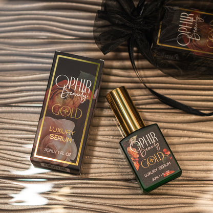 overhead view of a bottle of Ophir Gold Argan oil serum beside its gilded retail box, lying on a wavy reflective bronze surface