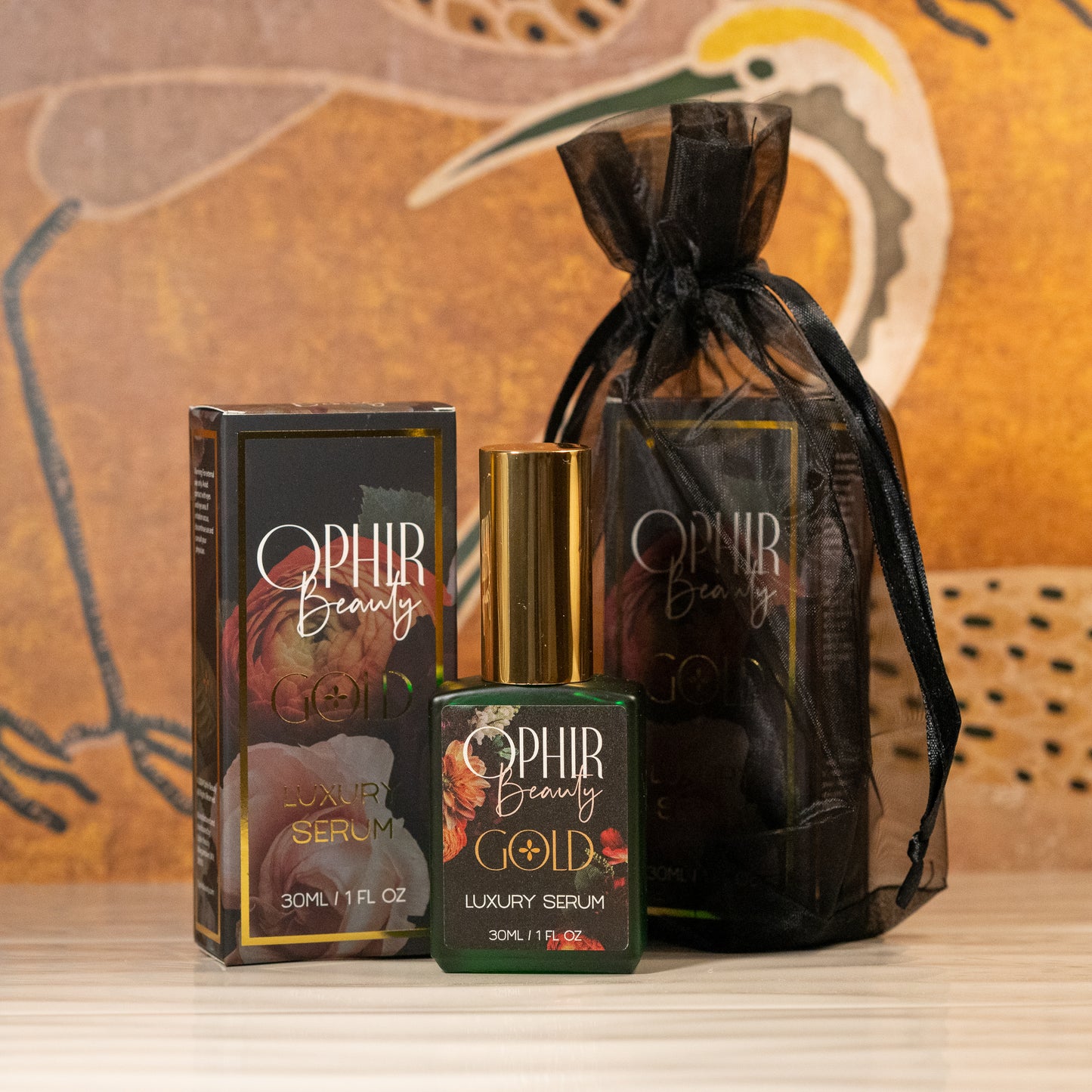 a green glass bottle of Ophir Gold with a black floral label stands beside its gilded retail box and black organza bag, on wavy white tiles with vintage wallpaper in the background