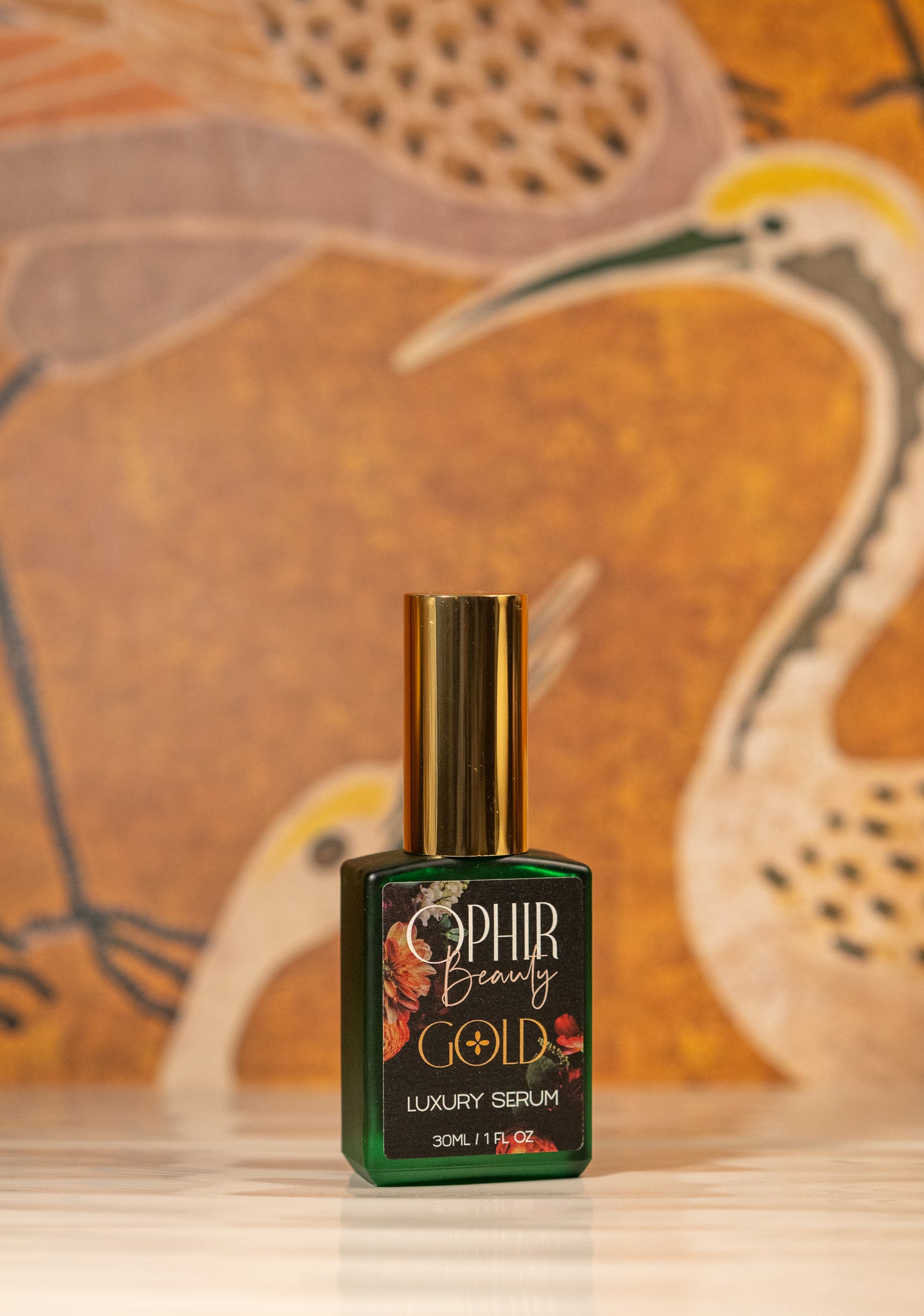 a green bottle of Ophir Gold Argan & Walnut oil Serum with a floral label sits on textured white tiles with a vintage gold crane wallpaper behind.