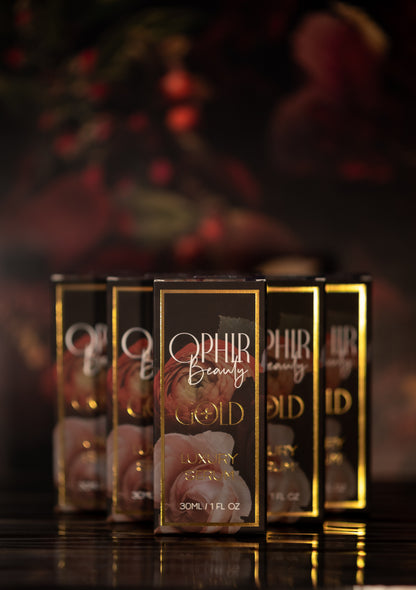 Several glimmering boxes of Ophir Gold Serum are arranged neatly in a pyramid on a dark reflective surface with hazy florals in the background.