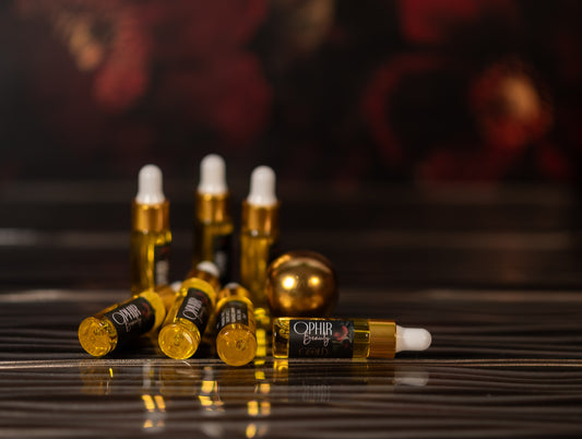 Several small glass dropper bottles filled with golden oil are scattered on a dark reflective glass with dark red florals in the background