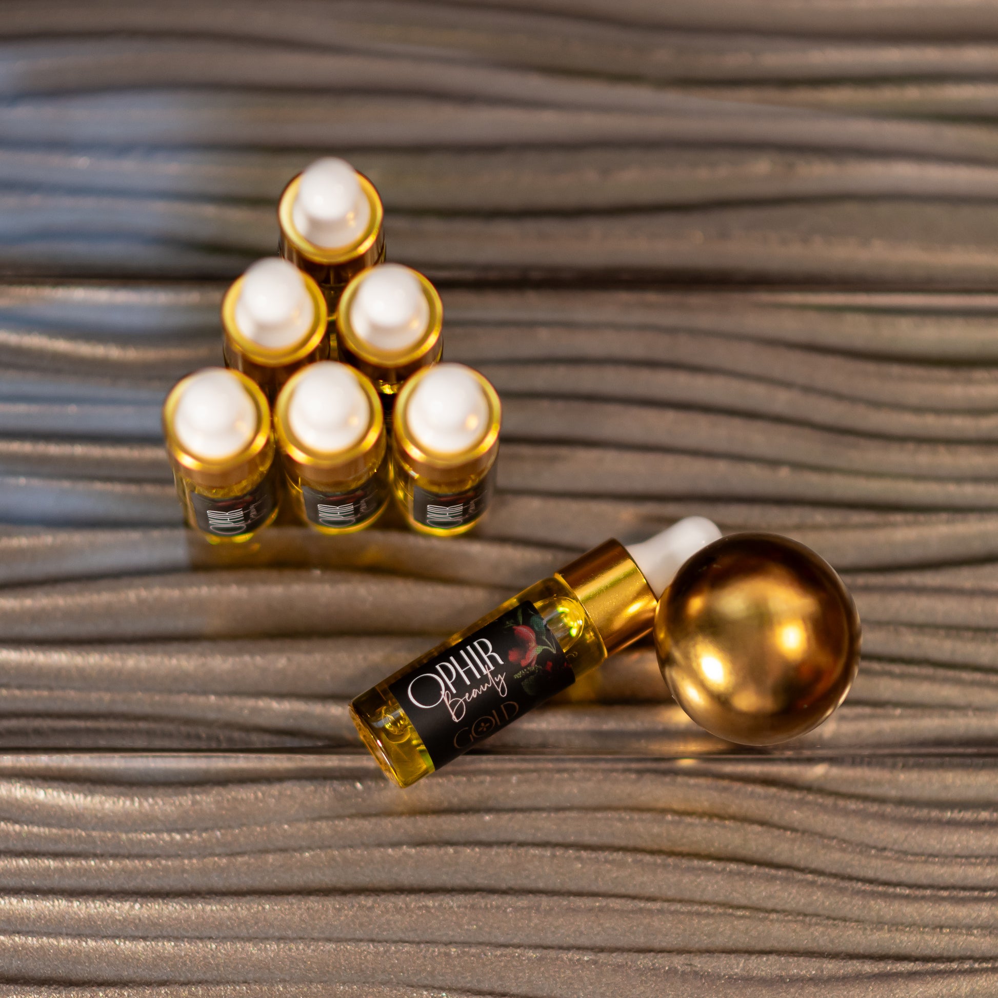 Overhead view of six small glass dropper bottles of Ophir Gold Serum arranged in a pyramid, with one bottle lying on its side, on a textured metallic bronze surface