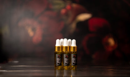 several small glass dropper bottles of Ophir Gold Luxury Serum stand neatly in a row on a dark surface, with hazy dark red florals in the background.