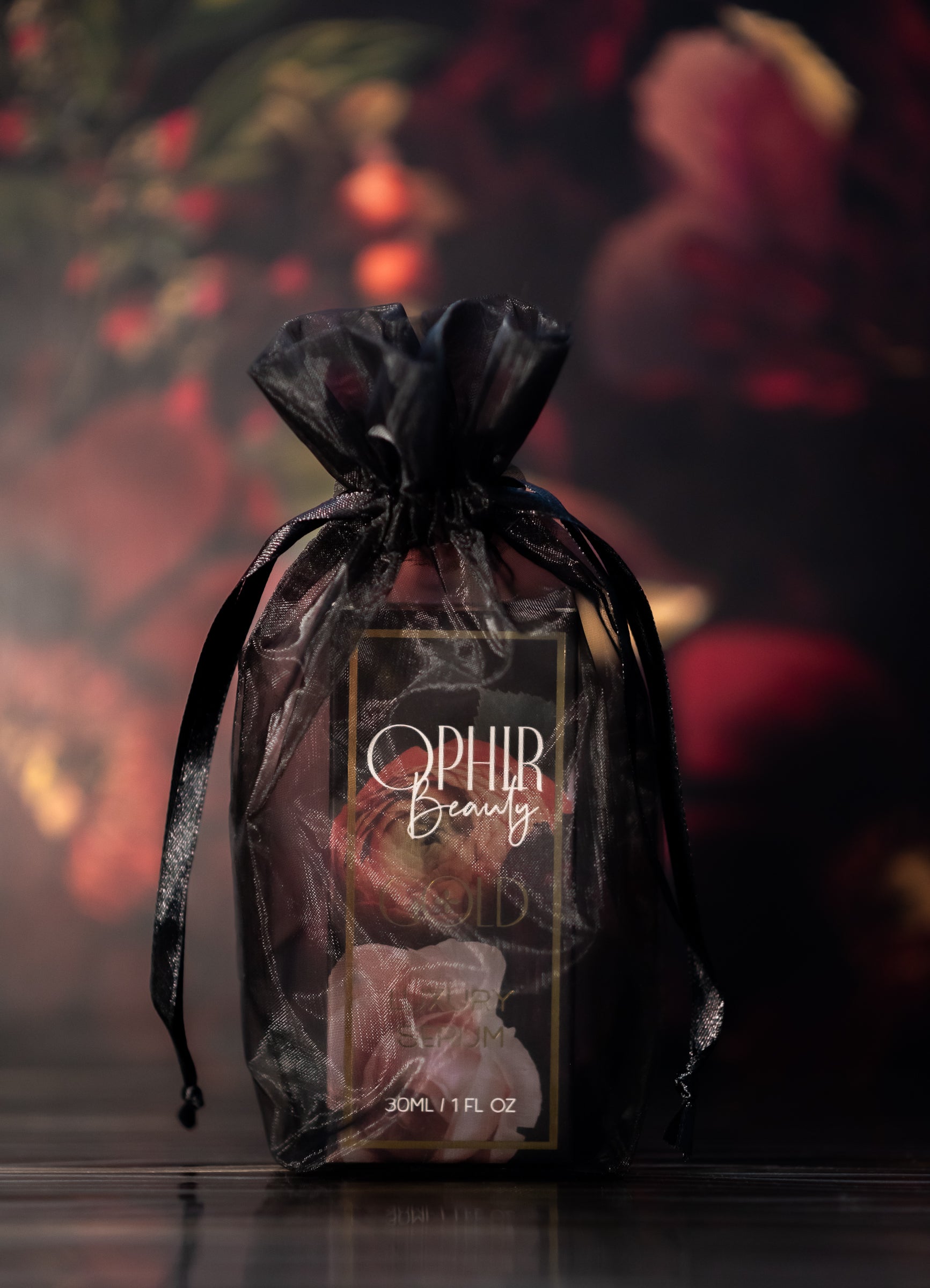 a black and floral box of Ophir Gold serum in its black organza bag stands on a dark reflective surface with florals in the background