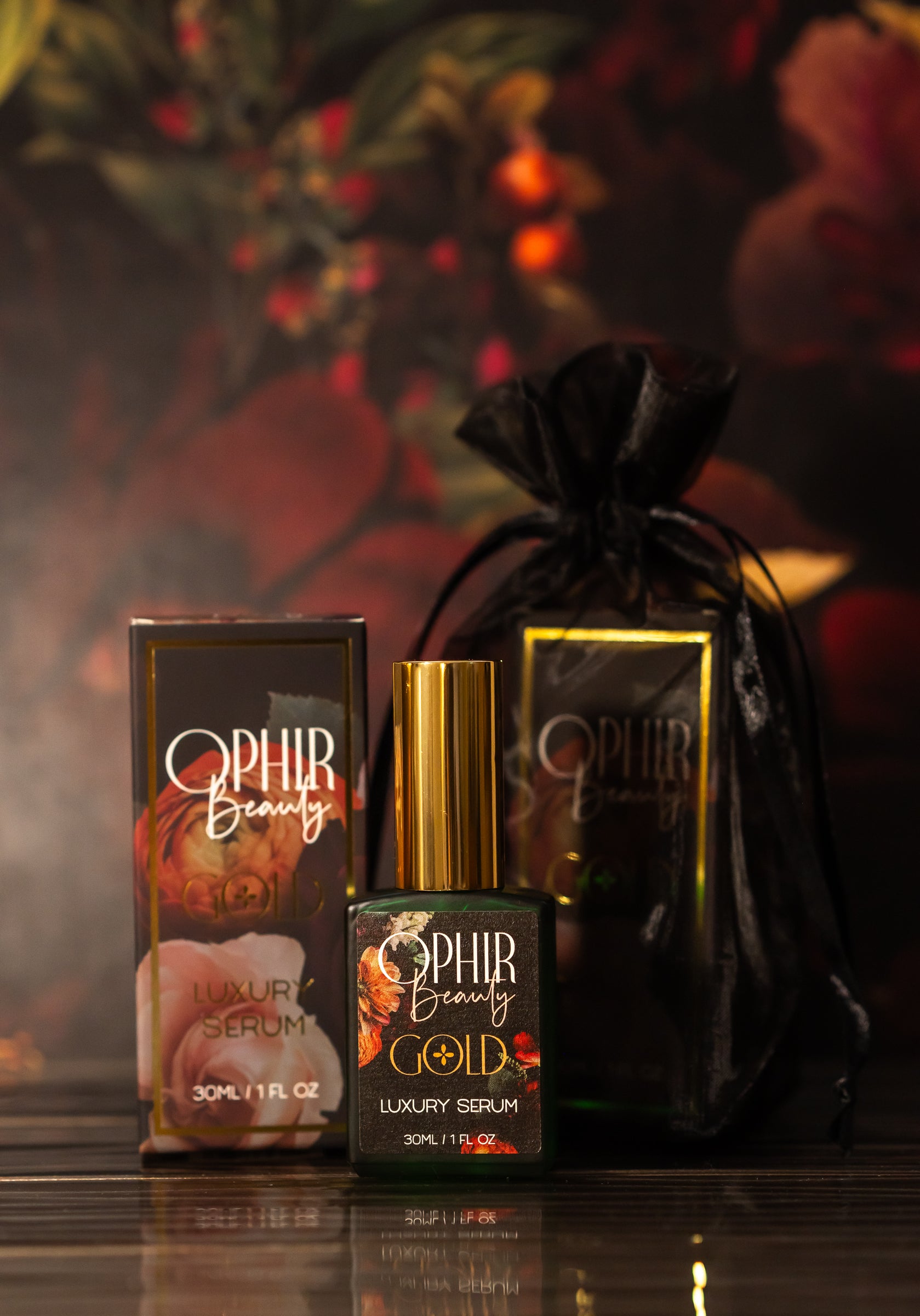 A green and metallic gold bottle of Ophir Gold Natural Luxury Argan oil Serum sits with its gilded floral box and black organza gift bag in front of a floral background